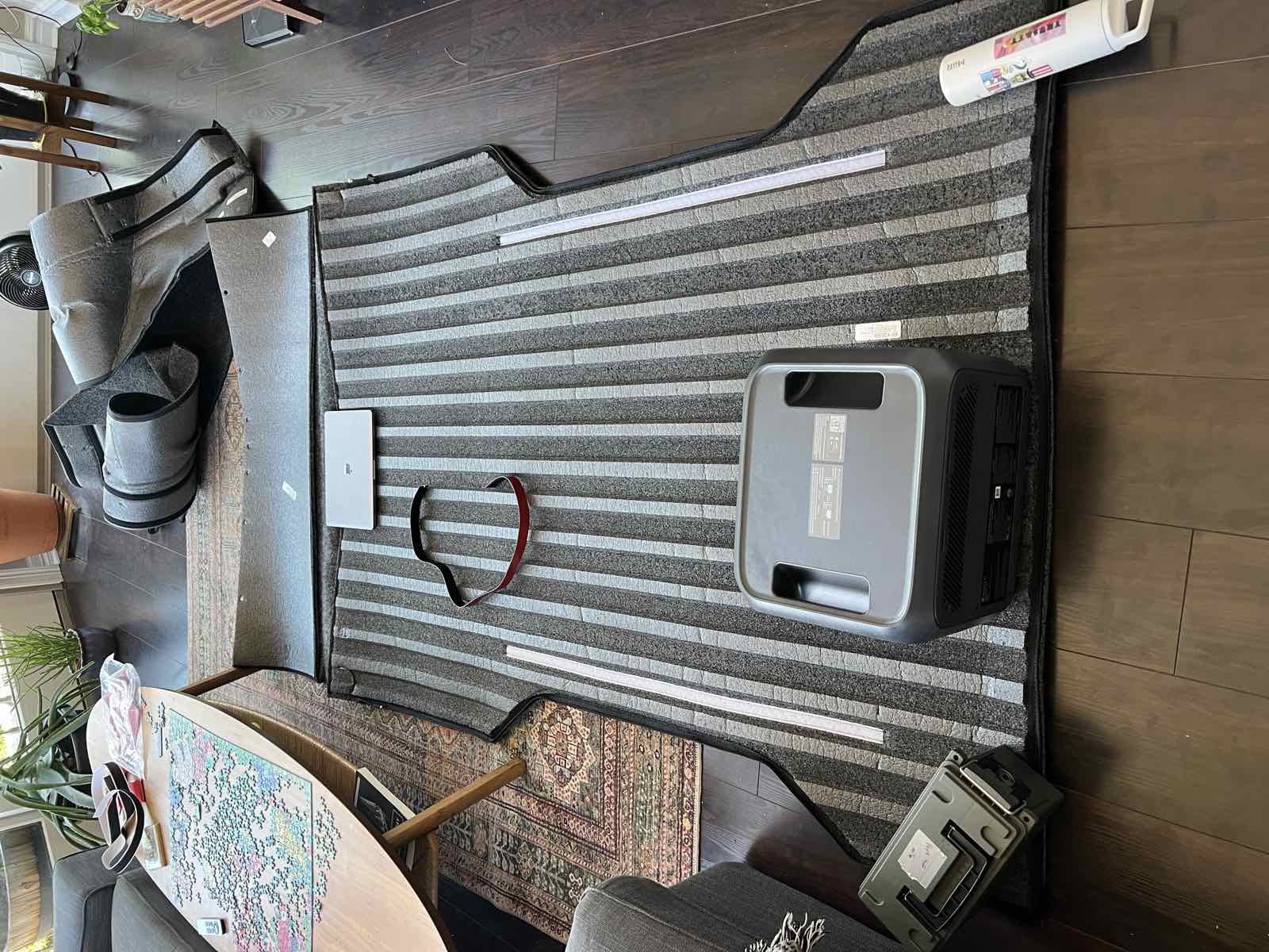 A Husky Guardian Comfort bed liner unfolded flat across a living room floor with a power station, a hard case and a laptop weighting it down, showing the sewn-in white velcro strips.