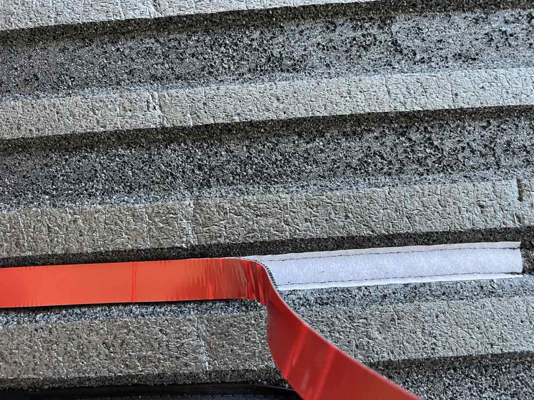 Close-up of a white velcro strip sewn into the underside of a bed liner with the red adhesive backing tape being peeled away.