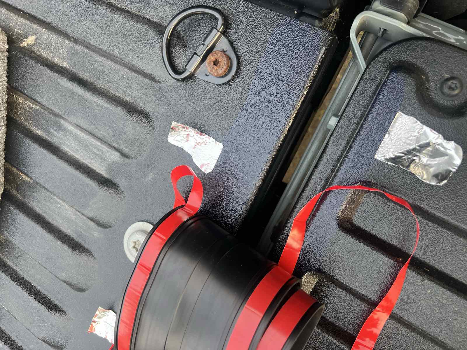 A coiled black rubber ROK Block tailgate gap cover with red 3M adhesive backing tape, resting in a spray-lined Tacoma bed next to a D-ring tie-down and small squares of aluminum tape.